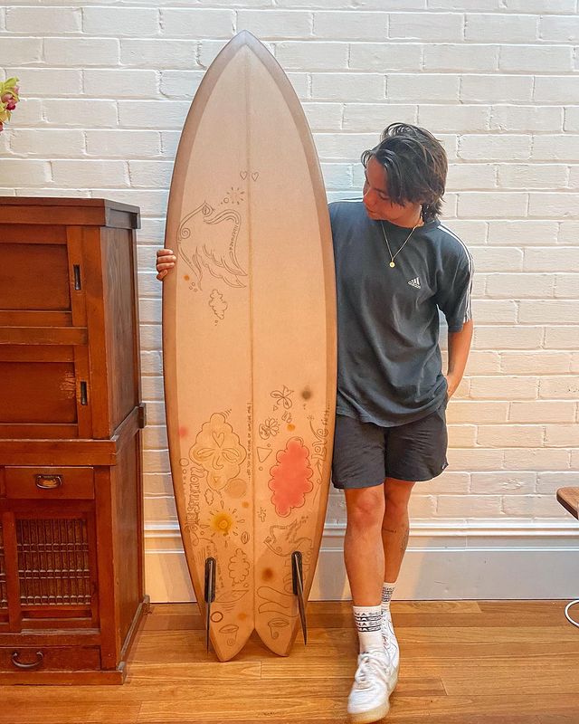 Zoe Terakes in a grey t-shirt and black shorts holding a new surf board.